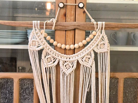 Natural cotton macramé wall hanging with an arc of wooden beads and long fringe, on driftwood
