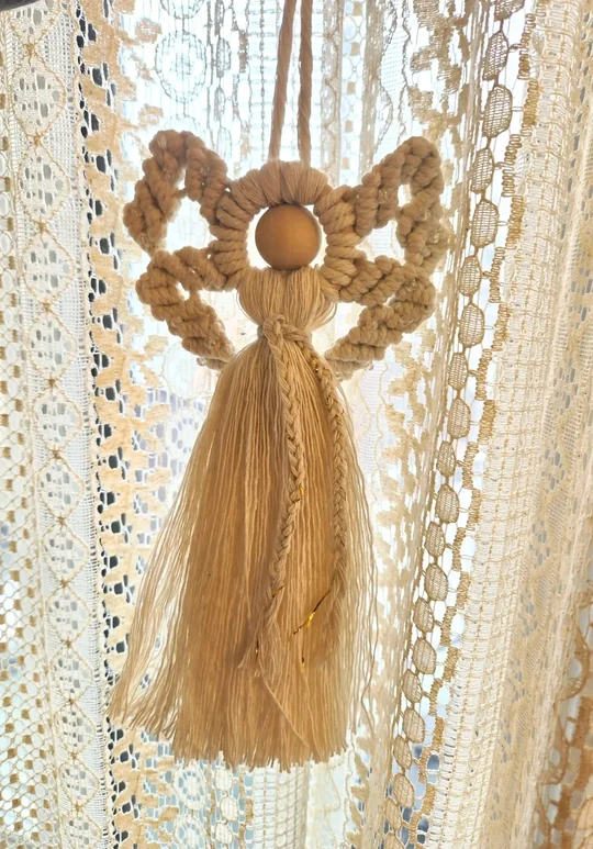 Caramel macramé angel with a long fringe skirt, in front of a lace curtain