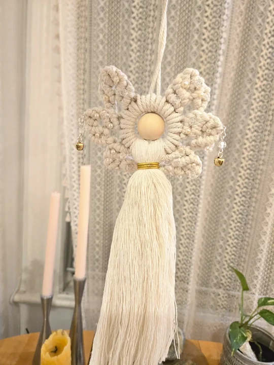 Cream macramé angel with bells beside candles and dried flowers