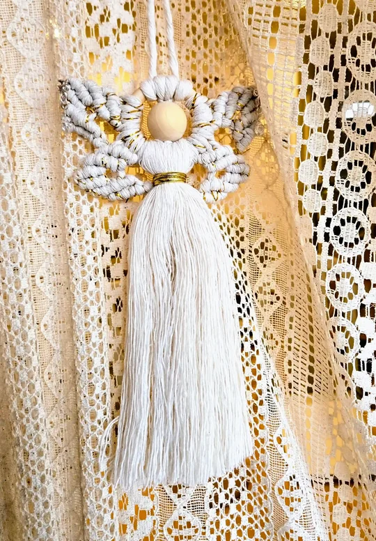 Cream macramé angel with curled wings and crystals, in front of a lace curtain